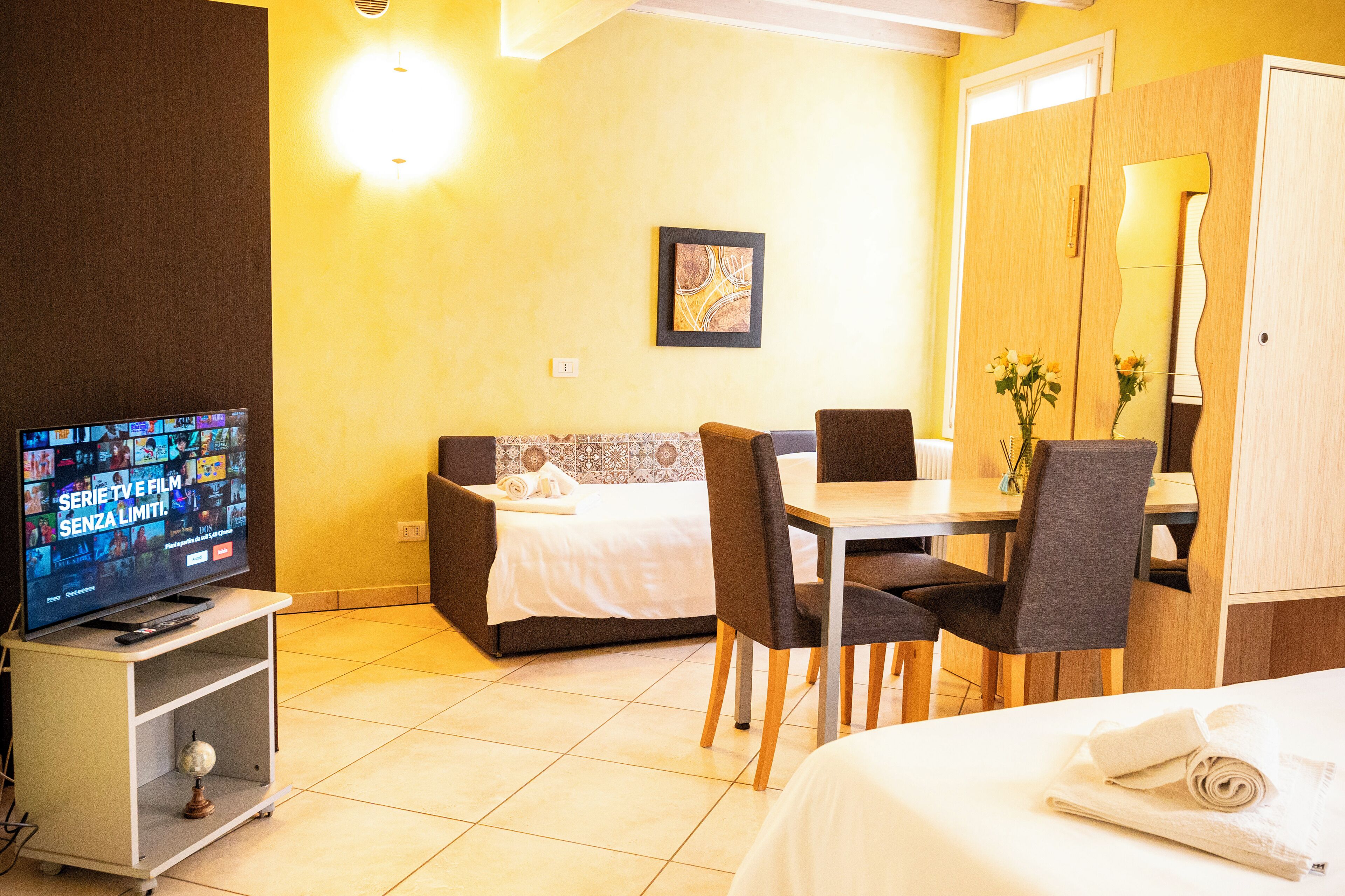 Quarto de hotel com cama, mesa de jantar com cadeiras, televisão sobre suporte e obra de arte pendurada na parede.