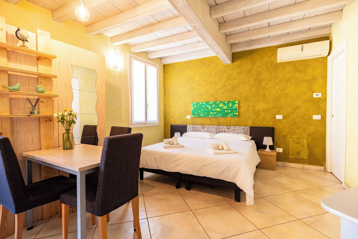 A hotel room with a bed, bedside table, lamp, and a dining area with chairs and a table.