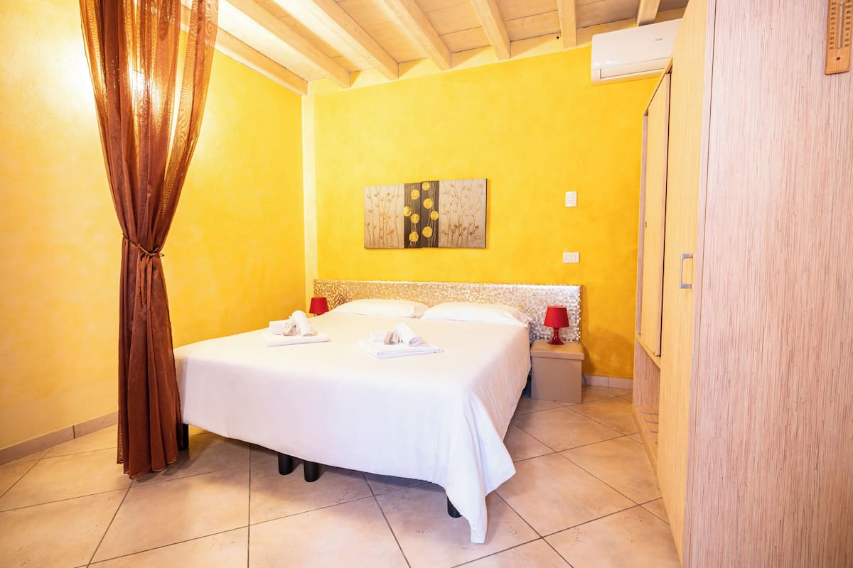 A hotel room with a bed, a painting on the wall, a red lamp, and a white air conditioning unit.