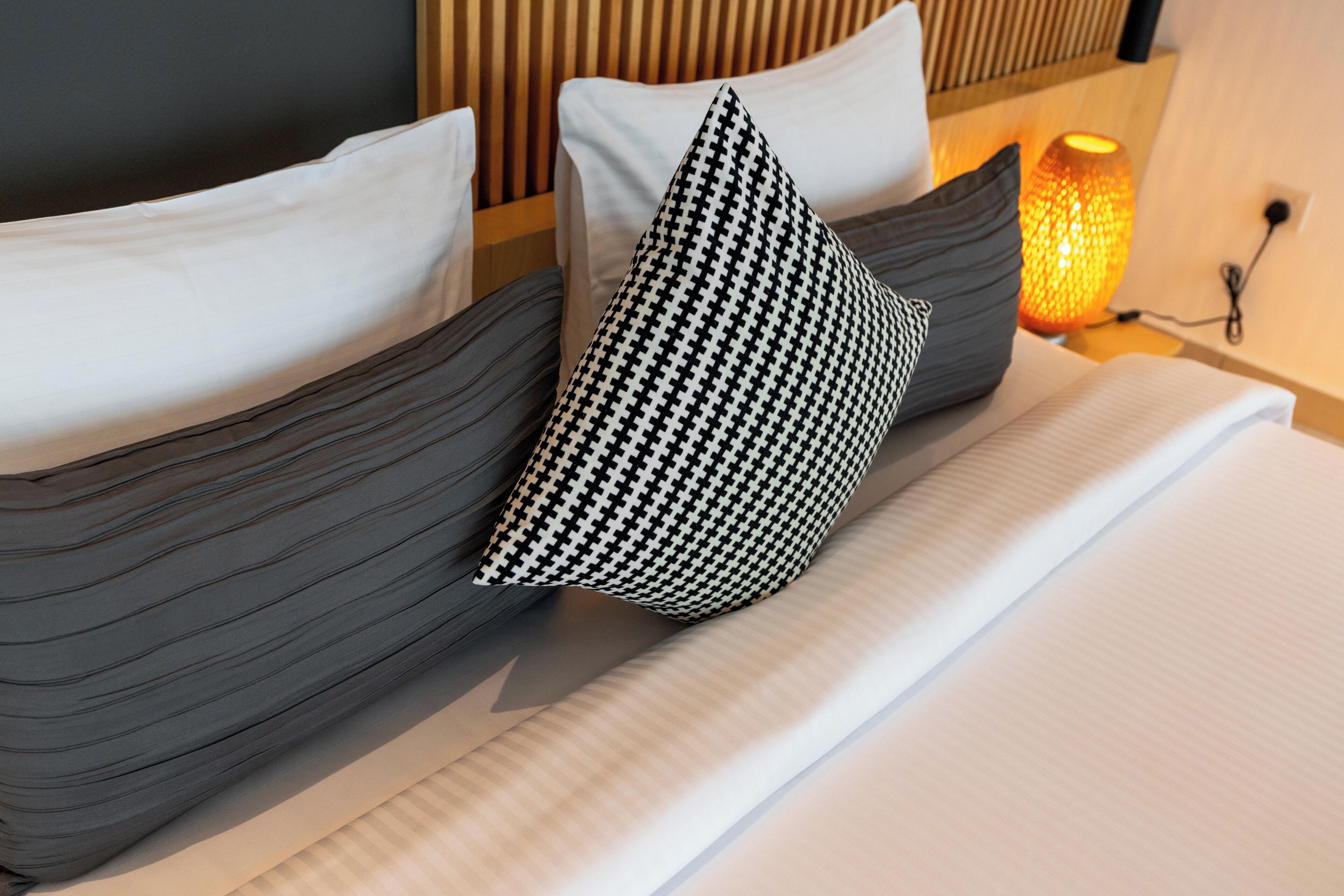 A neatly made bed with striped pillows and a houndstooth pillow, a bedside lamp, and a power outlet on the wall.
