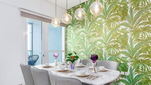 A dining area with a round table set for four, grey upholstered chairs, a tropical leaf-patterned wall, and hanging pendant lights. - Samaya Holiday Homes - Marina luxury Suites (Dubai)