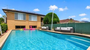 Una piscina con una casa in mattoni sullo sfondo, una recinzione verde e un albero.