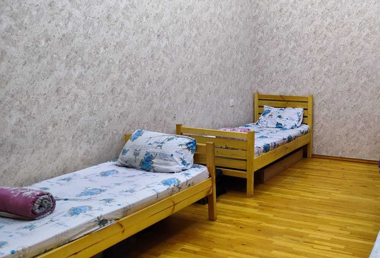 A small room with two wooden beds, each with a floral-patterned mattress and pillows. The room has a wooden floor and a textured wall.