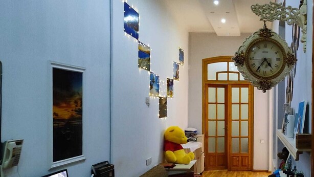Una habitació amb sòl de fusta, una finestra gran, un escriptori amb un ordinador, un os de peluix i un rellotge de paret.
