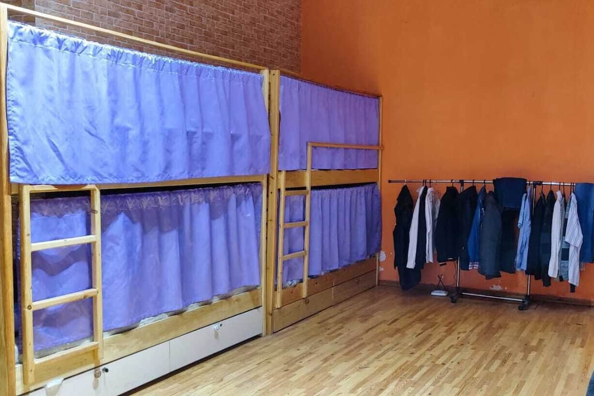 Quarto com duas beliches cobertas por cortinas roxas, um cabideiro com diversas peças de roupa e uma parede de tijolos.