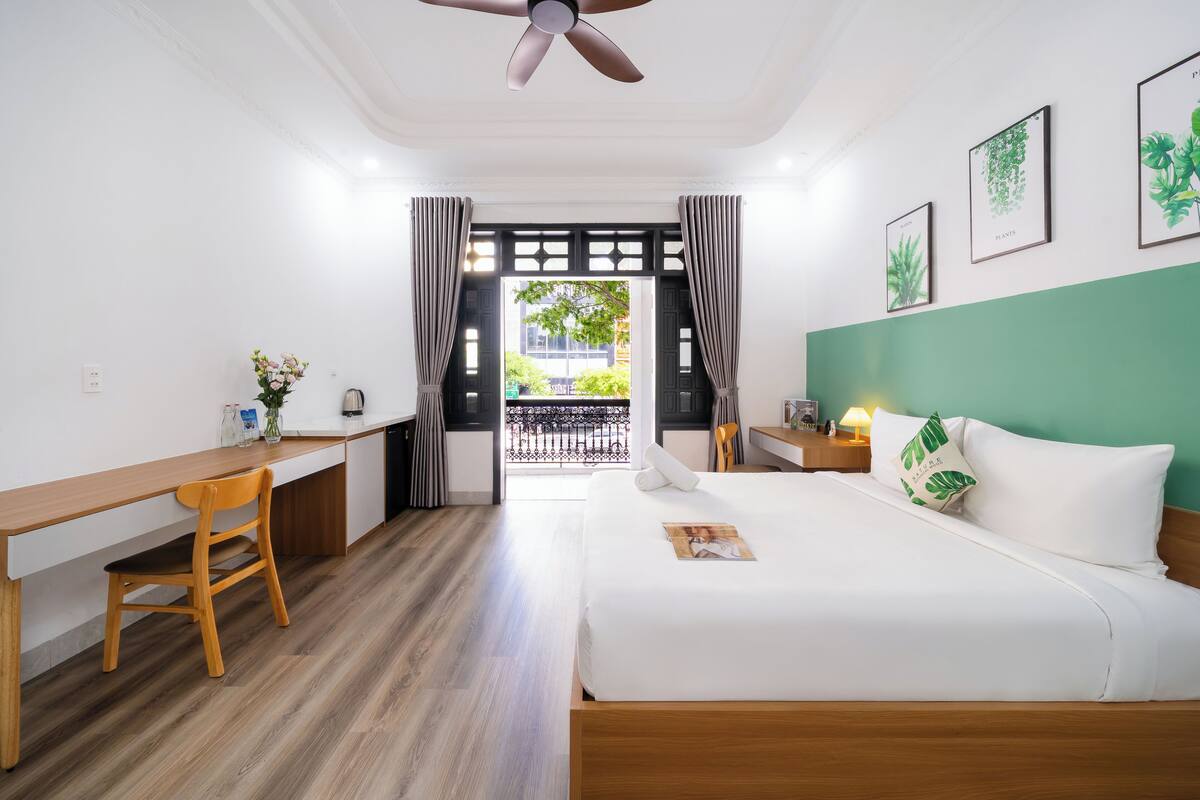 A hotel room with a wooden desk, a bed with white linens, a ceiling fan, and a view through a window with curtains.