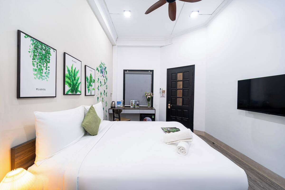 A modern hotel room with a neatly made bed, a desk with a computer, a flat-screen TV, and framed botanical artwork on the wall.