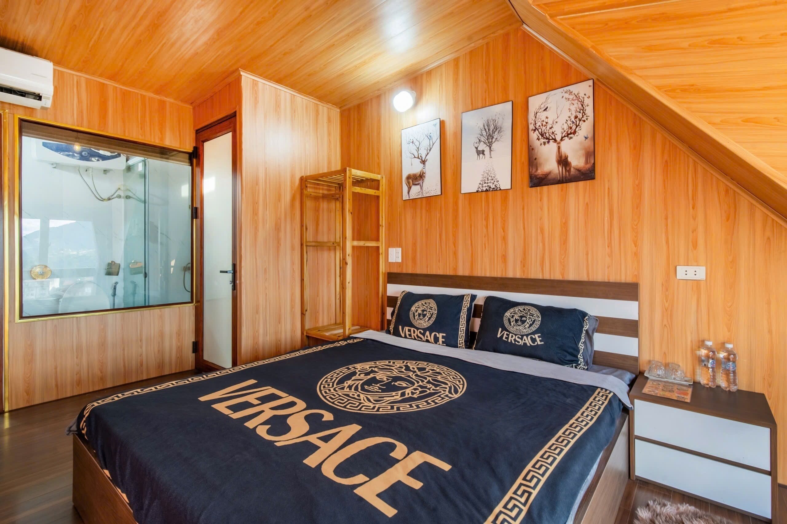 A wooden-paneled bedroom with a bed featuring a 'Versace' branded bedspread, a wooden nightstand, and a glass shower enclosure.