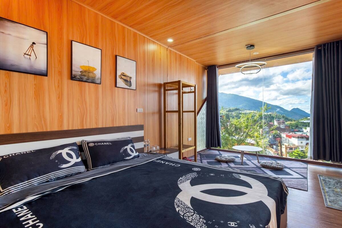 A hotel room with wooden paneling, a bed with Chanel bedding, a balcony with a view of mountains and a town, and artwork on the walls.