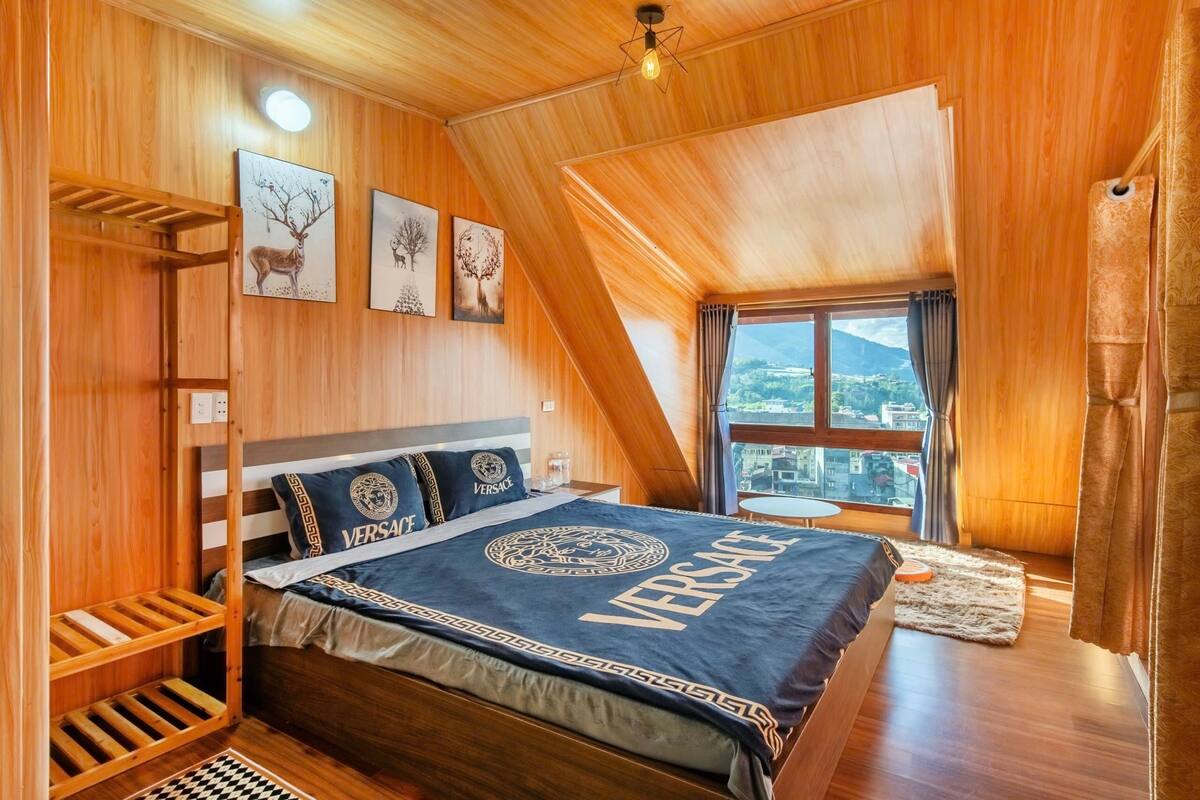 A bedroom with a sloped ceiling, wooden walls, a bed with a 'Versace' blanket, a window with a view, and a shelf with decorative items.