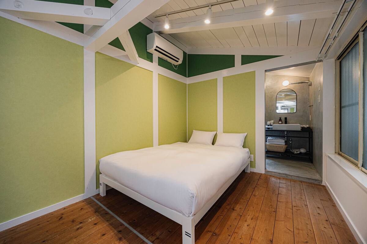 A bedroom with a bed, wooden flooring, green and white walls, a window, and a bathroom visible through an open door.