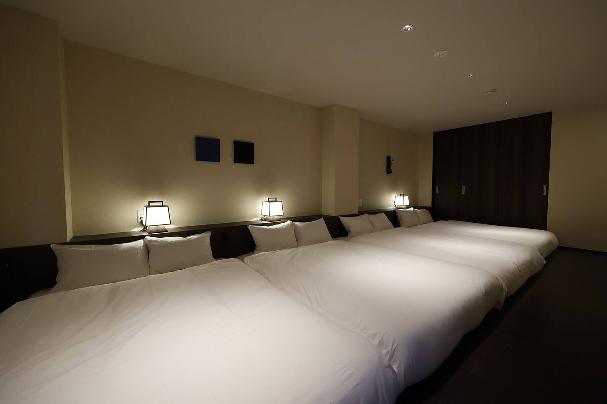 A hotel room with two beds, each with a nightstand and a lamp. The room has a dark wooden wardrobe and beige walls.