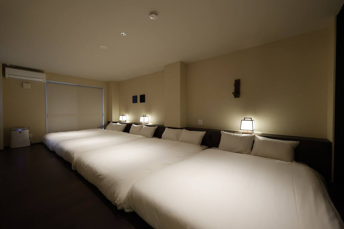 A hotel room with three single beds aligned in a row, a bedside table, a wall-mounted lamp, and a ceiling light.