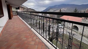 Ein Balkon mit Fliesenboden und Metallgeländer, der auf eine gebirgige Landschaft mit Gebäuden und einer Stebrücke blickt.