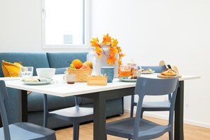 Un comedor con una mesa blanca, sillas azules y un sofá azul. Se ven platos de comida, un jarrón con hojas anaranjadas y un vaso de jugo de naranja.