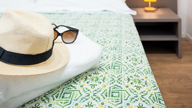Una cama bien hecha con una colcha estampada, dos almohadas anaranjadas brillantes, una lámpara de noche, un sombrero de paja y un par de gafas sobre la cama.