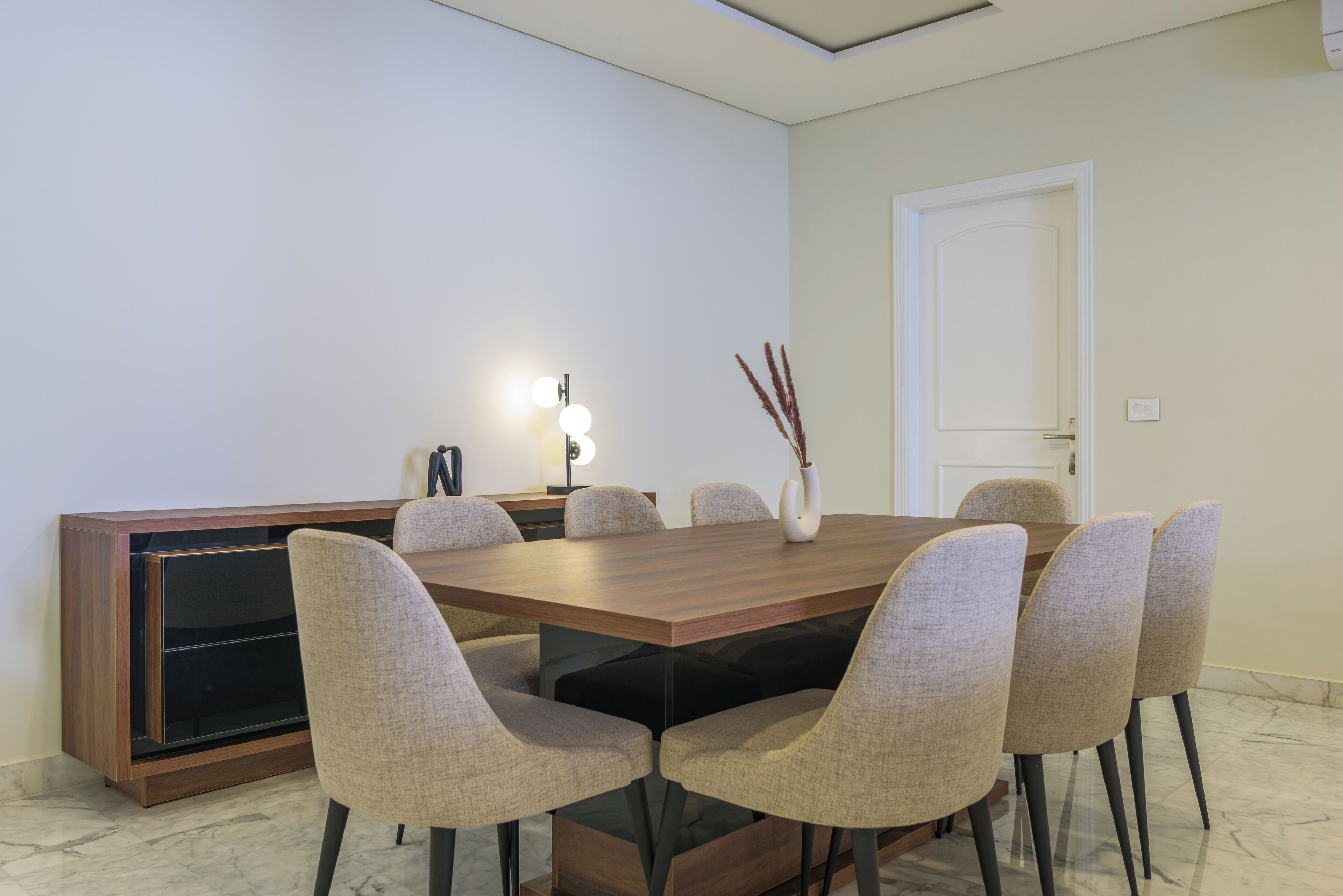 A modern dining room with a wooden table, six upholstered chairs, a vase with decorative branches, and a wall-mounted light fixture.