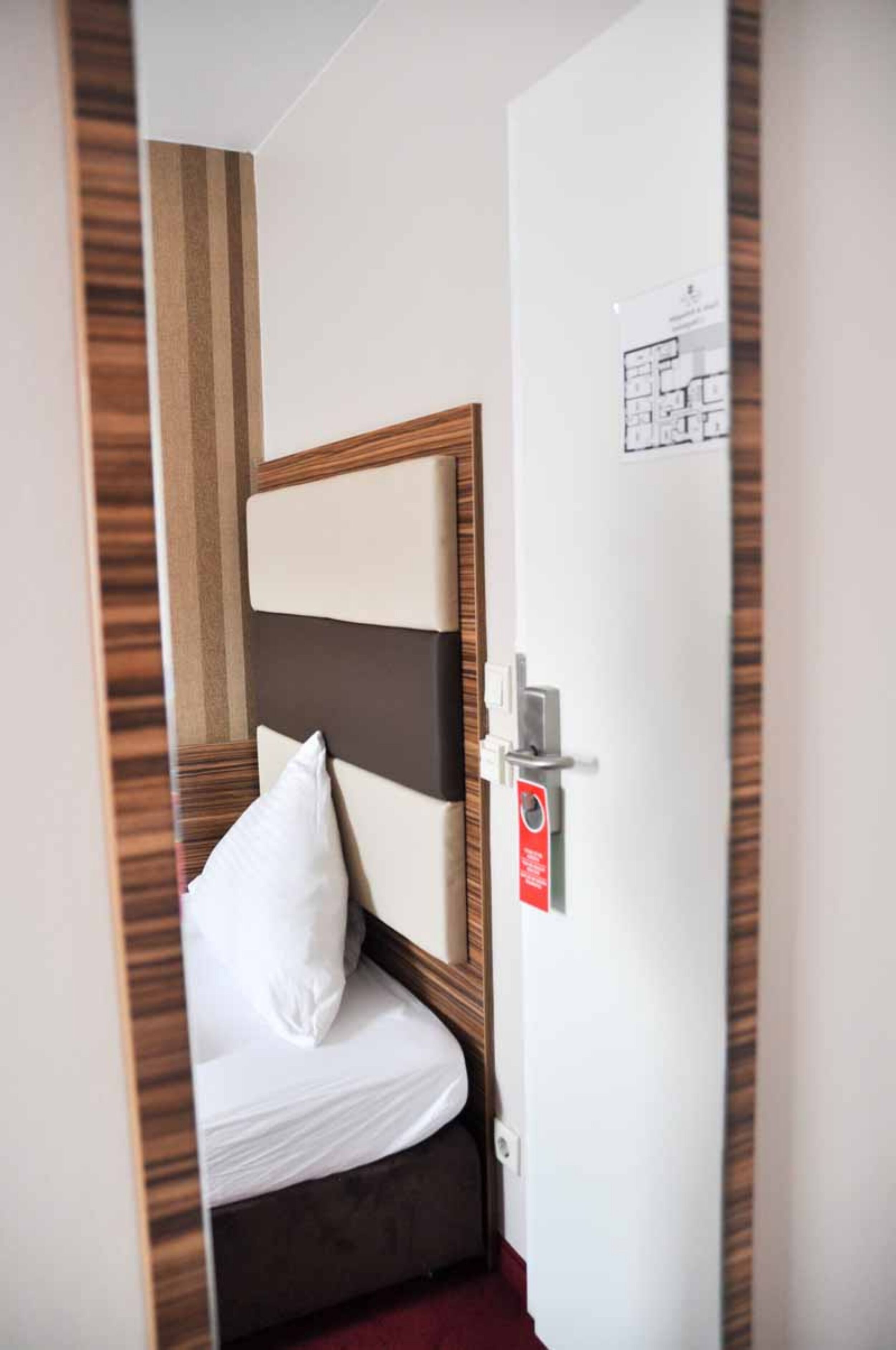 A hotel room with a bed, a door with a red emergency exit sign, and a wall-mounted light switch.