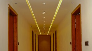 Um corredor comprido e estreito, com portas de madeira em ambos os lados, piso brilhante e iluminação embutida com detalhes amarelos.