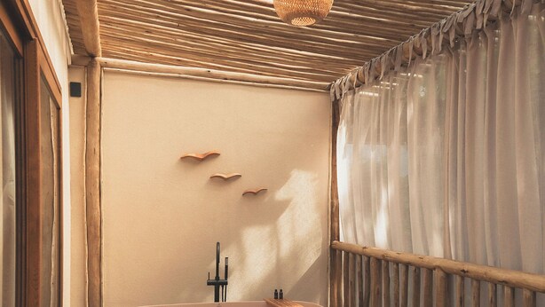 Una habitación con una mesa redonda, sillas de mimbre, un sofá con cojines, una bañera y un techo de listones de madera con una lámpara colgante.