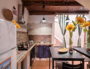 A kitchen with a brick wall, wooden ceiling, and hanging pendant lights. There is a dining table with a vase of sunflowers and a bowl. - Vista Sul Blu Private Residence 3 (Porto Cesareo)