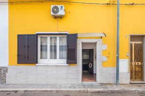 A building with a yellow exterior, a window with shutters, an air conditioning unit, and a door with a decorative design. - Vista Sul Blu Private Residence 3 (Porto Cesareo)