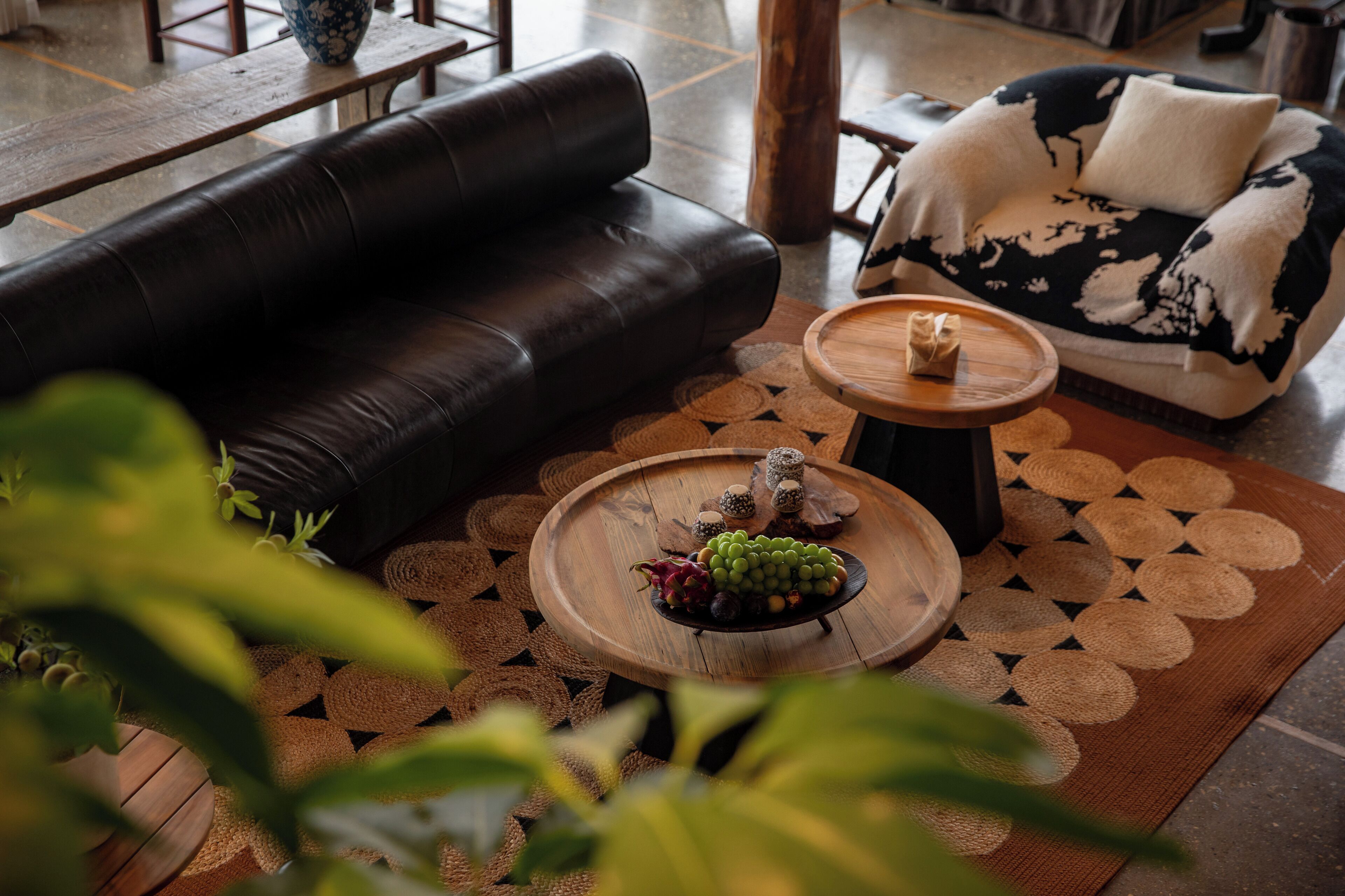 A modern living room with a black leather sofa, a patterned armchair, and a wooden coffee table with fruit and decorative items.
