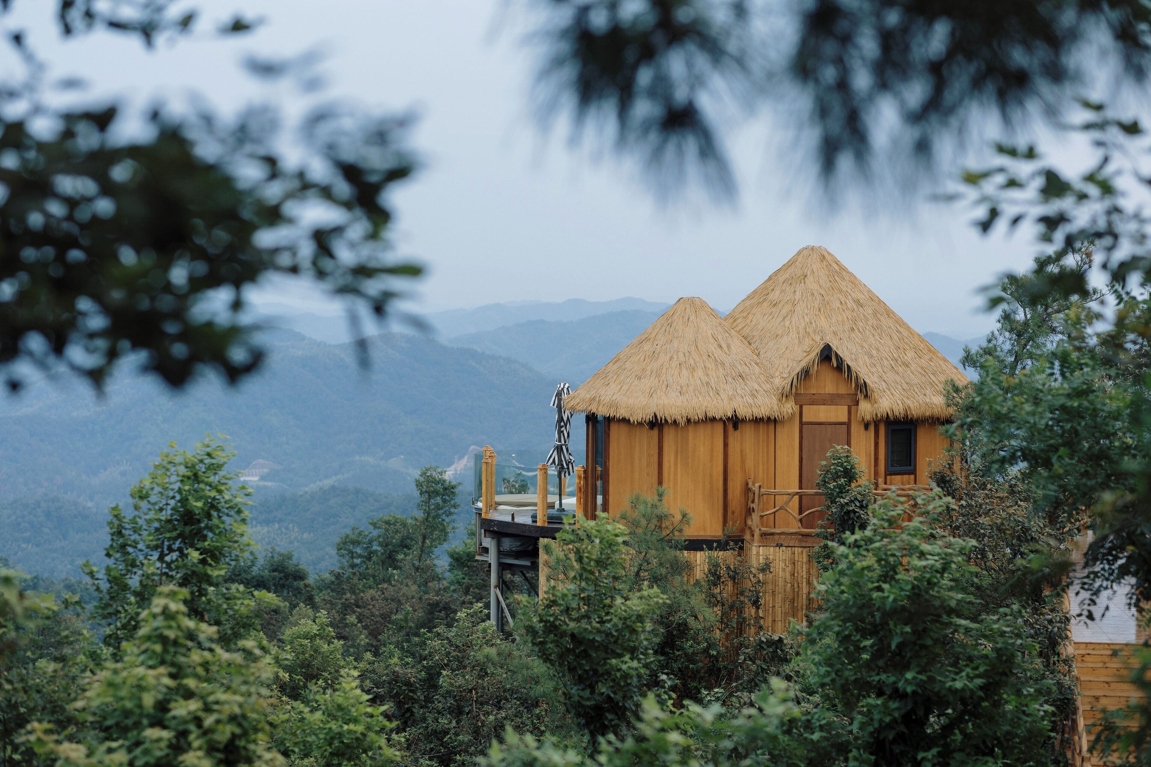 A wooden cabin with a thatched roof, surrounded by lush greenery and mountains in the background.