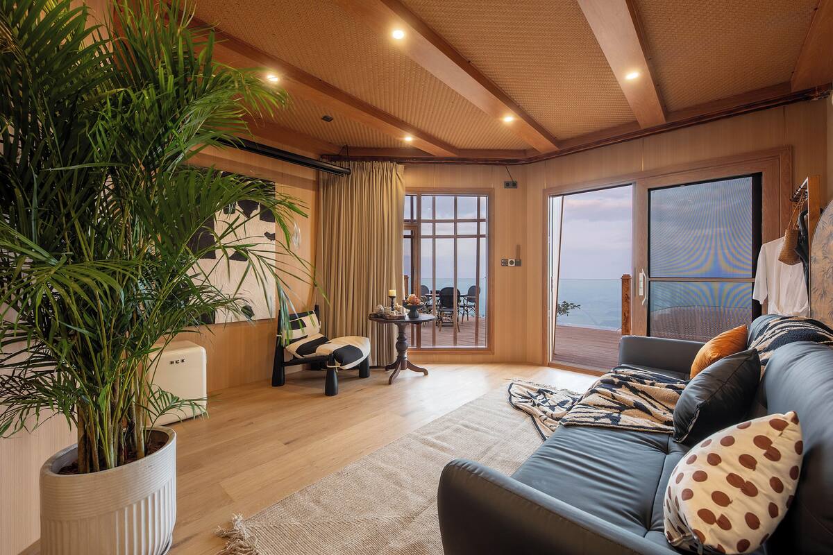 Una sala de estar moderna con una planta grande en maceta, un sofá con cojines estampados, un comedor con mesa y sillas, y vistas a través de un ventanal amplio.