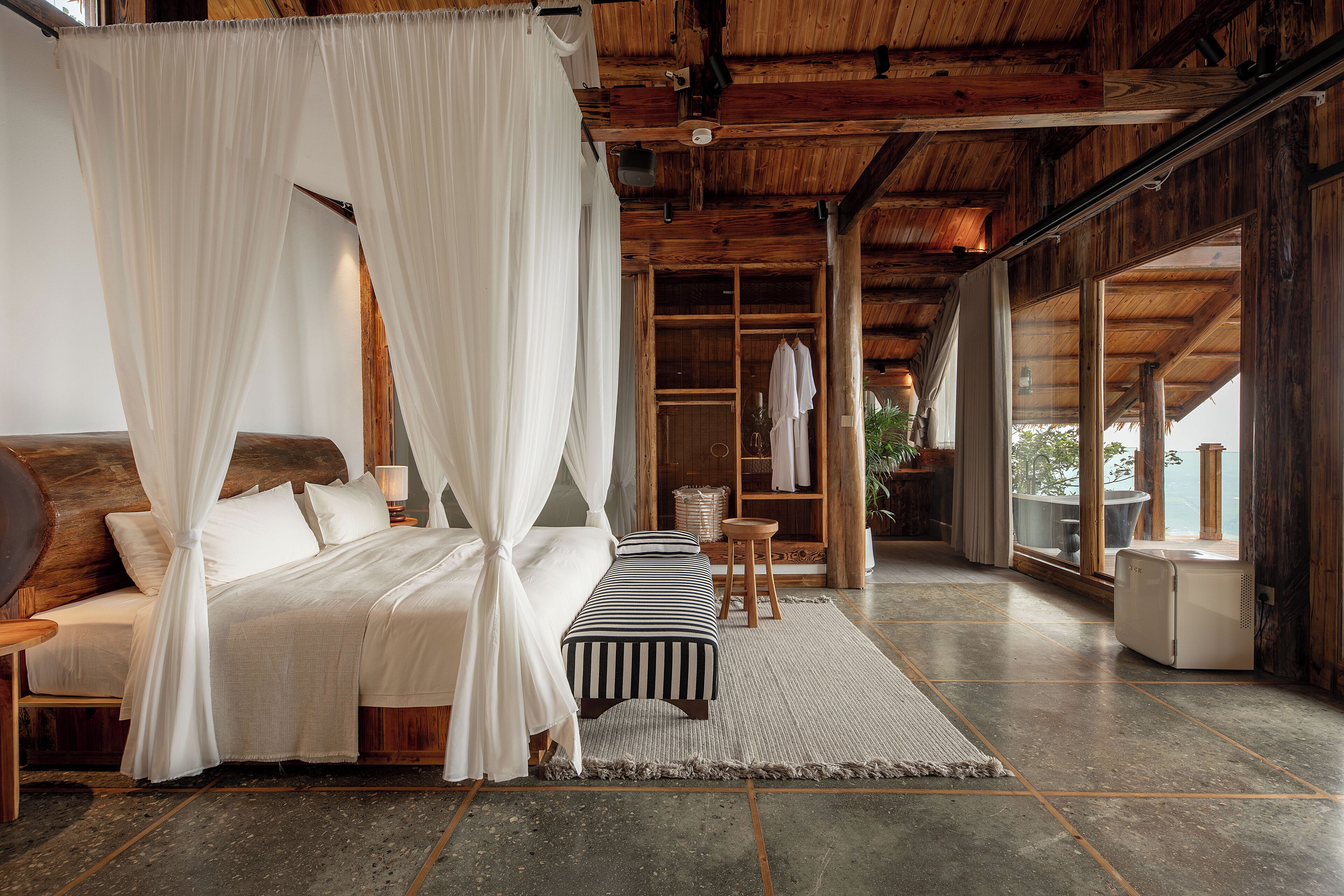 A bedroom with a canopy bed, a bedside table, a bench, and a large window.