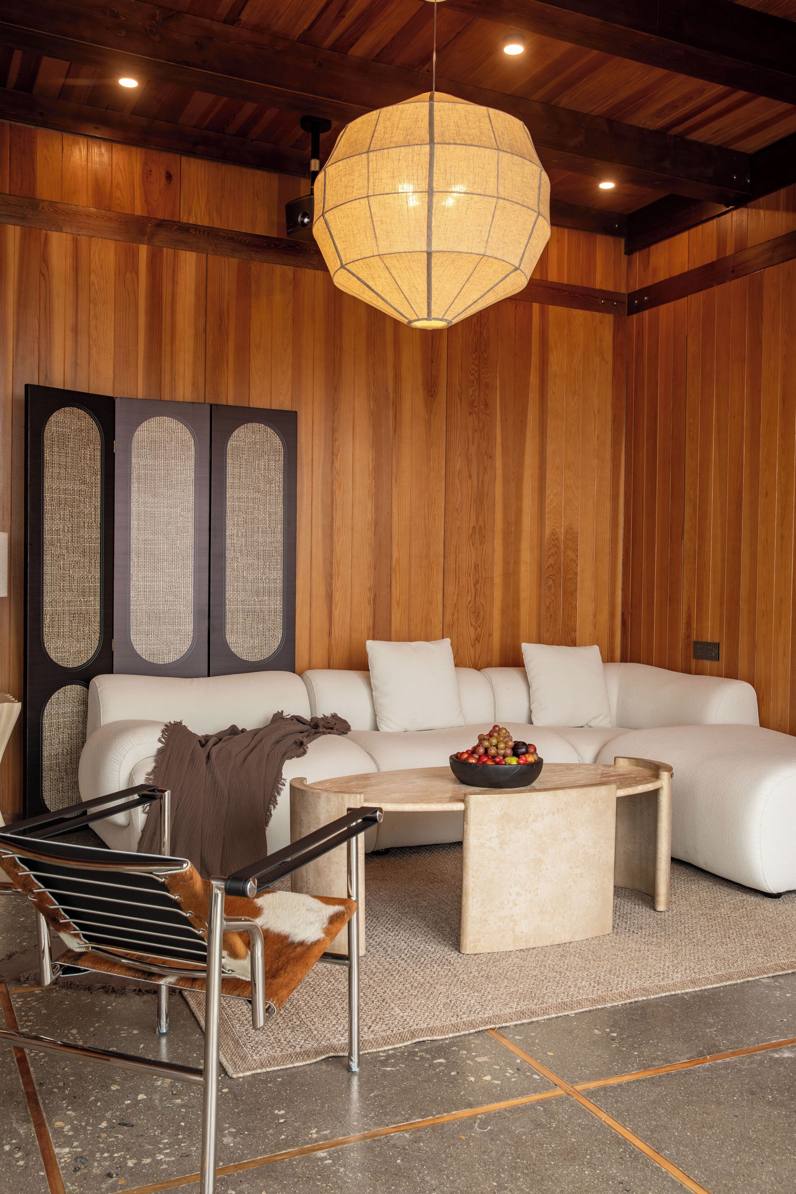 A modern living room with a white sofa, a wooden accent wall, a hanging light fixture, and a coffee table with a bowl of fruit.