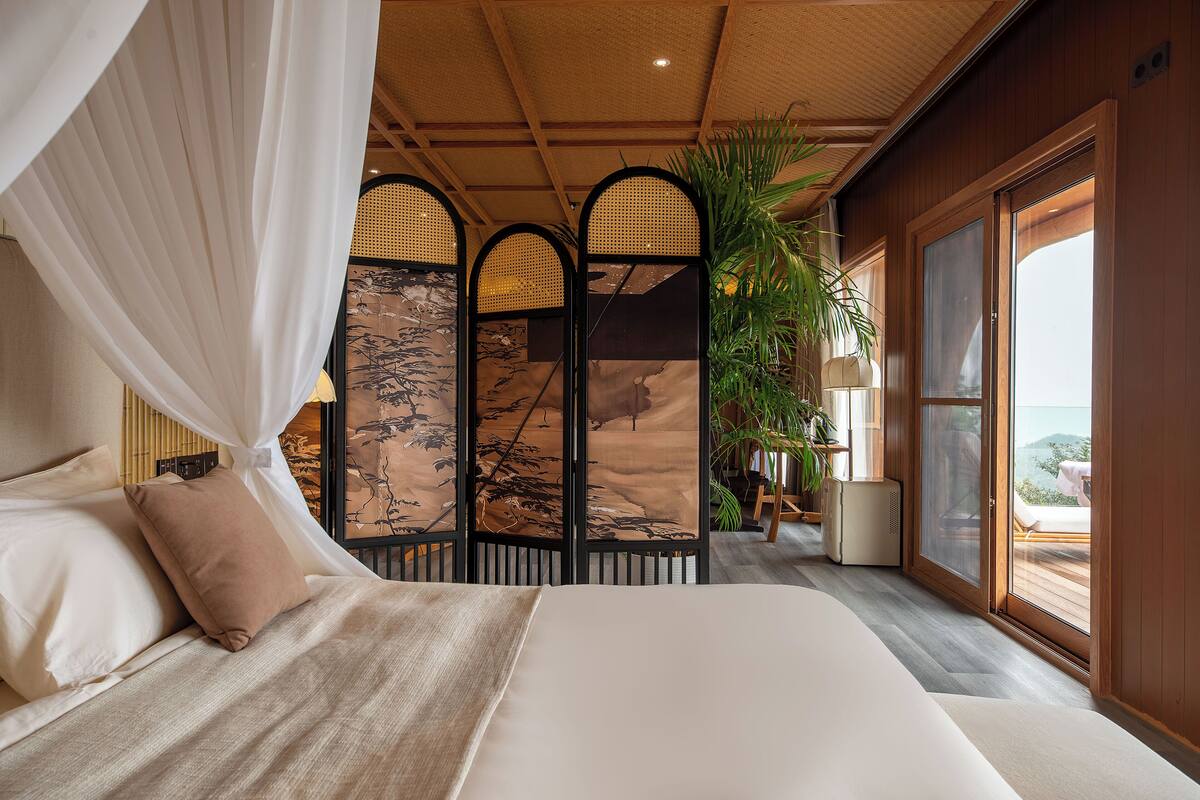 Un dormitorio con una cama grande, dosel, puerta corredera a un balcón y un biombo decorativo con una planta.