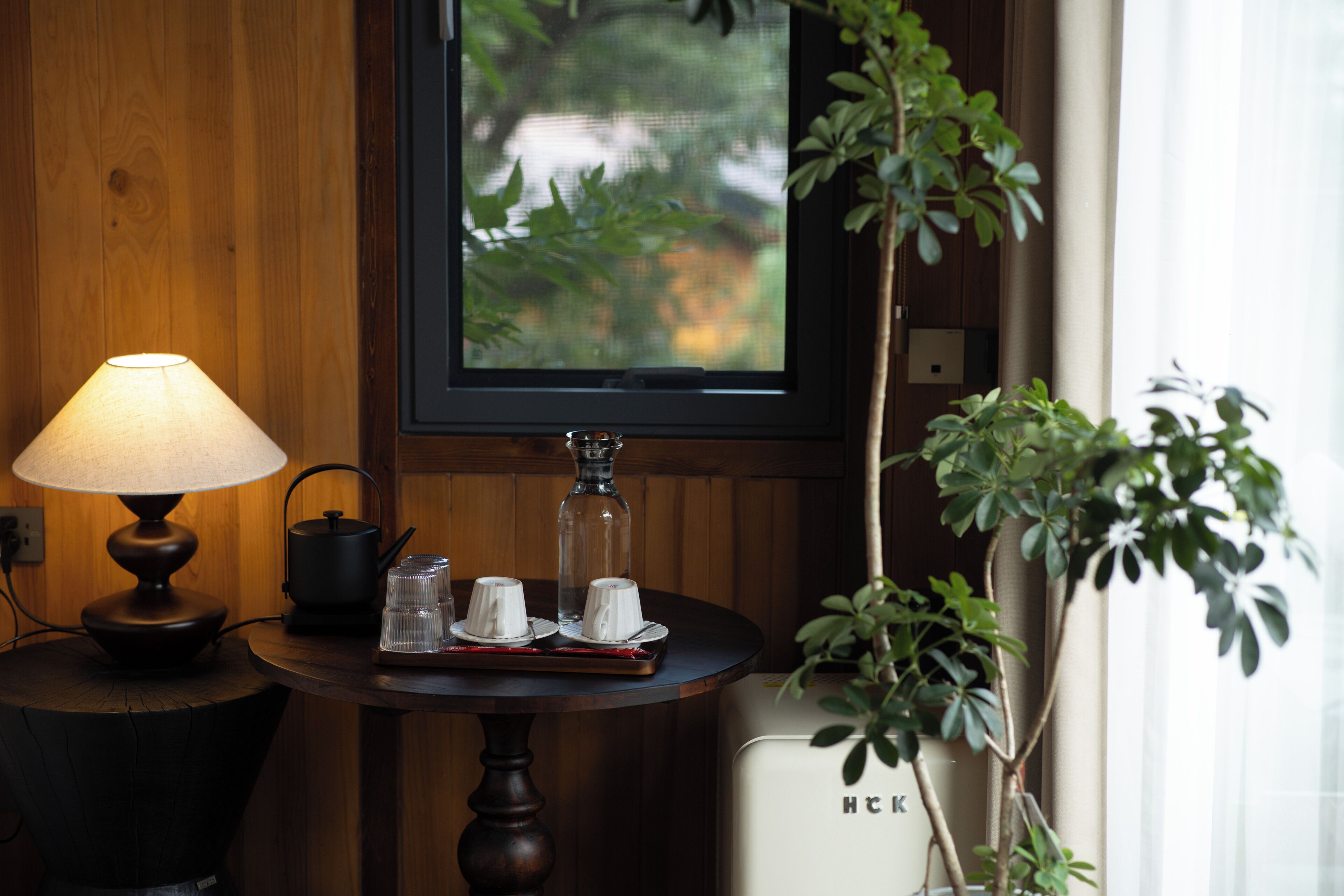 A wooden wall, a table with a lamp, a window with a view, and a plant.