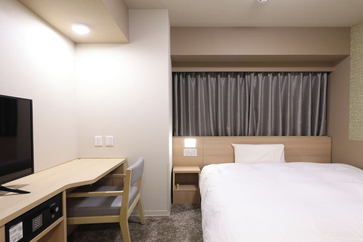 Habitación de hotel con cama, escritorio con televisor, silla, mesita de noche y ventana con cortinas.