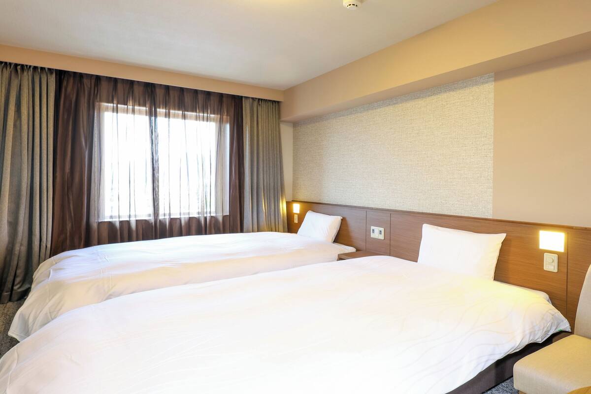 Habitación de hotel con dos camas, un ventanal con cortinas y una lámpara fijada a la pared.