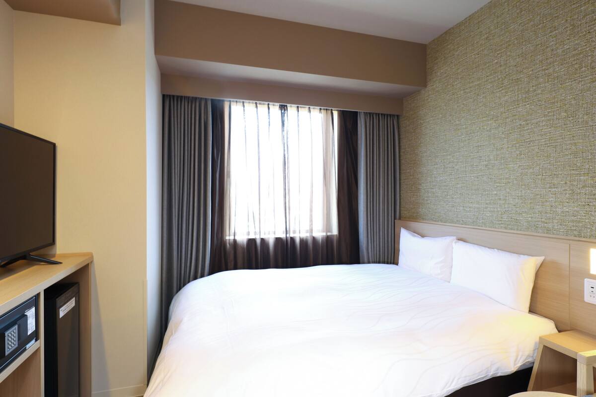 Habitación de hotel con una cama grande, un televisor sobre un mueble y una ventana con cortinas.