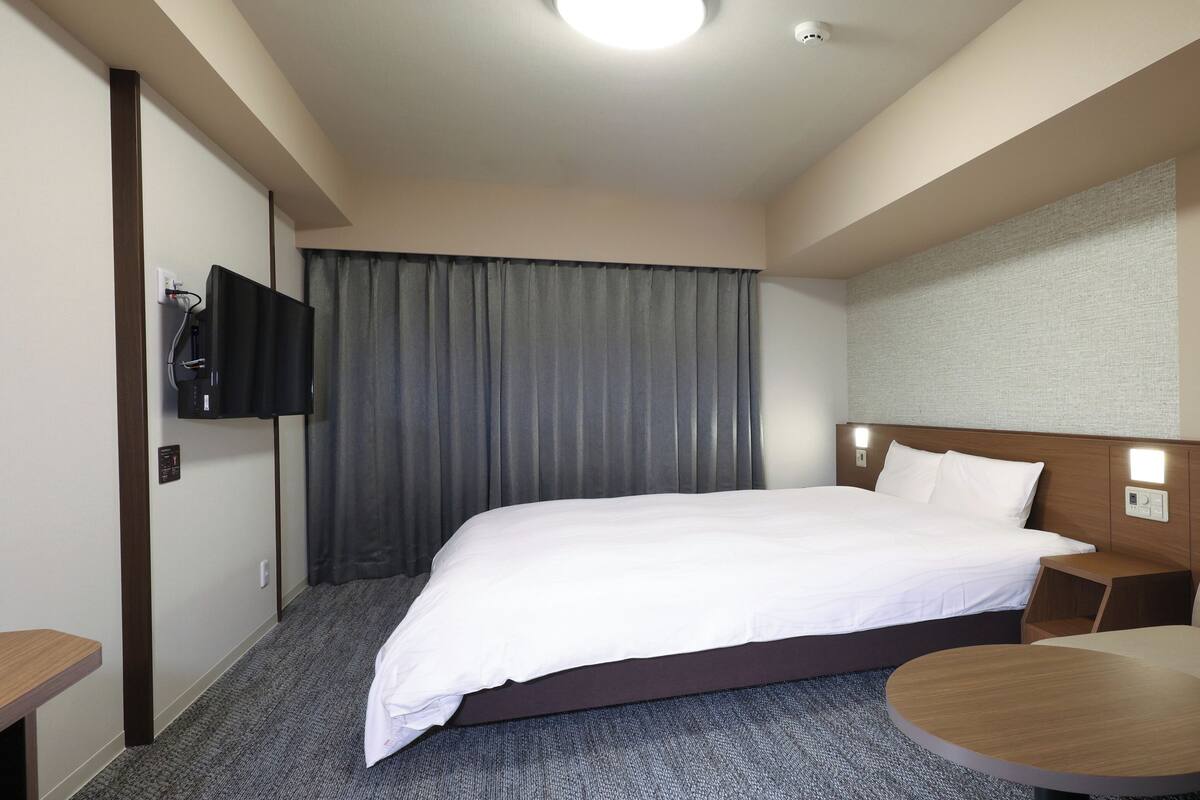 Una habitación de hotel con cama, sofá, una mesita, televisor montado en la pared y ventana con cortinas.