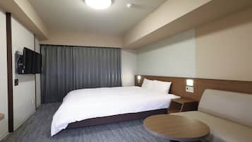 A hotel room with a bed, a sofa, a small table, a TV mounted on the wall, and a window with curtains.