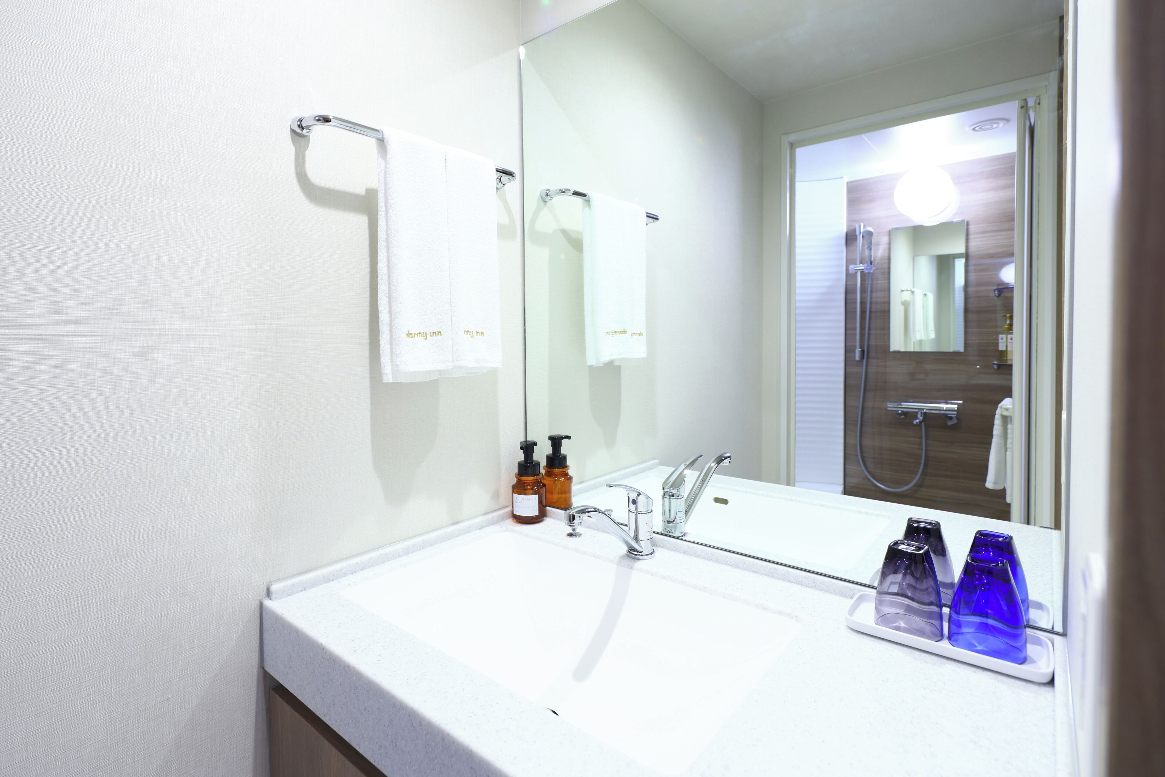 A bathroom with a white sink, two soap dispensers, a mirror, and a shower visible in the background.