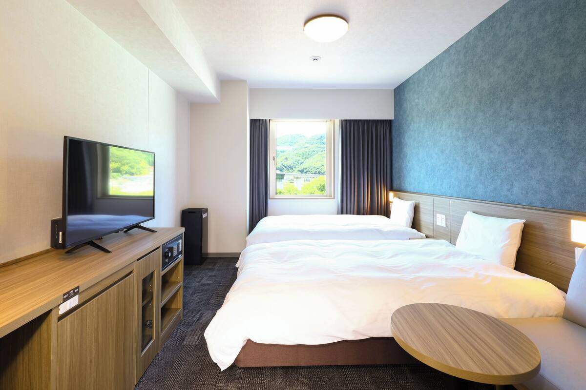 A hotel room with a bed, a TV, a small table, and a window with curtains.