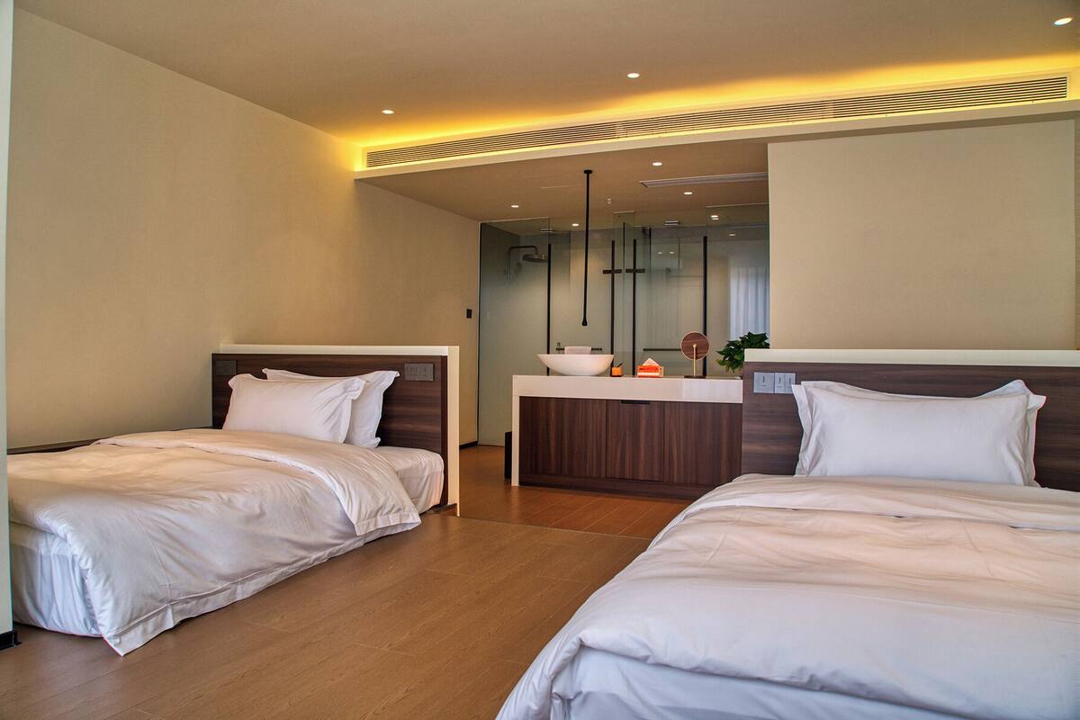 Une chambre d’hôtel avec deux lits simples, une petite salle de bain avec un lavabo et un miroir, et un design moderne avec un revêtement de sol en bois et un éclairage encastré.