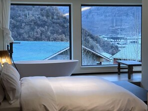 Une chambre moderne avec un grand lit, une table de chevet et une vue sur un paysage enneigé, visible à travers de grandes fenêtres.