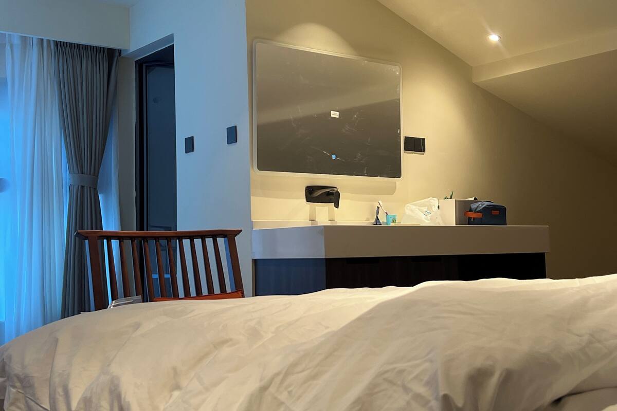 Une chambre d’hôtel avec un lit, une chaise en bois, un bureau avec un miroir et une fenêtre avec des rideaux.