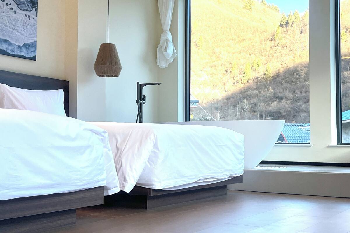 Une chambre moderne avec un grand lit, une table de chevet, une lampe fixée au mur et une fenêtre donnant sur une colline boisée.