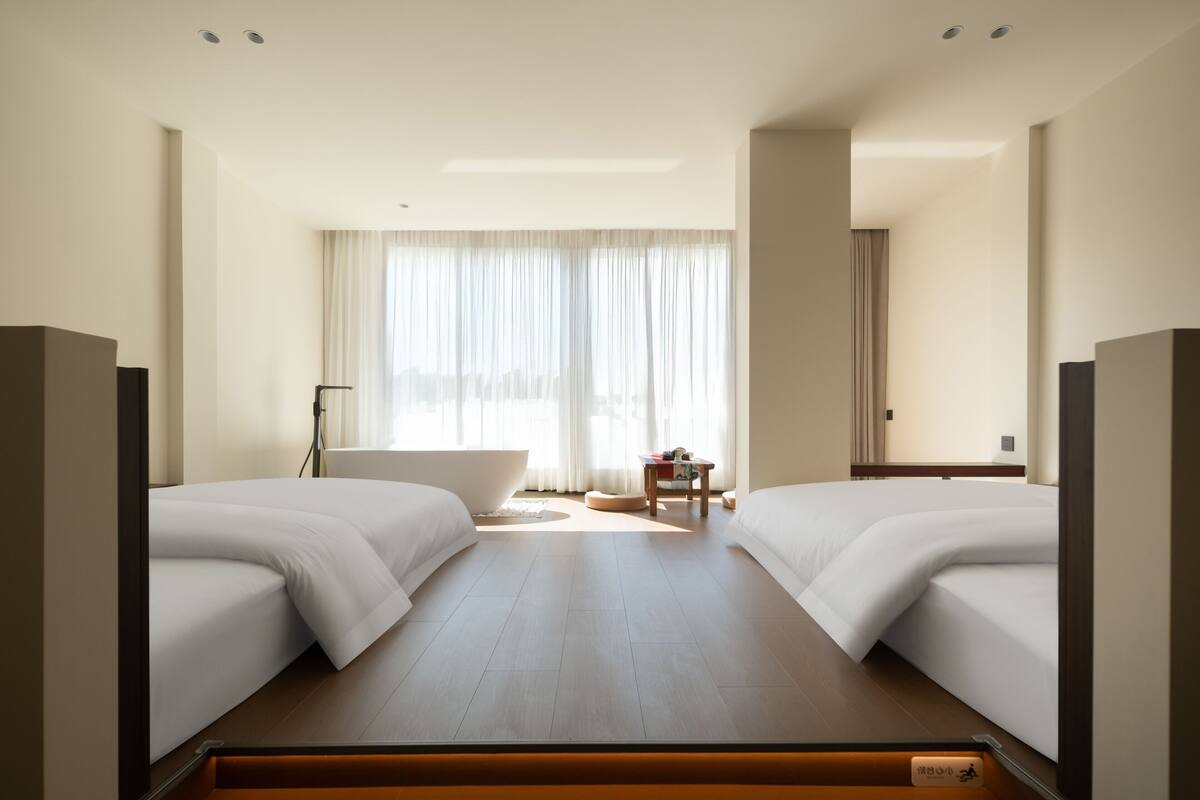 Une chambre d’hôtel avec deux lits, une baignoire, une petite table et une grande fenêtre avec des rideaux.