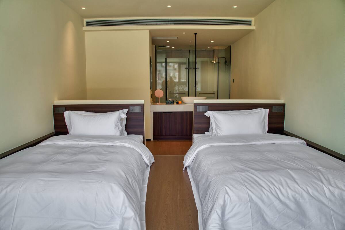 Une chambre d’hôtel avec deux lits, une salle de bain avec une douche en verre, et une petite table avec un miroir et un bol.
