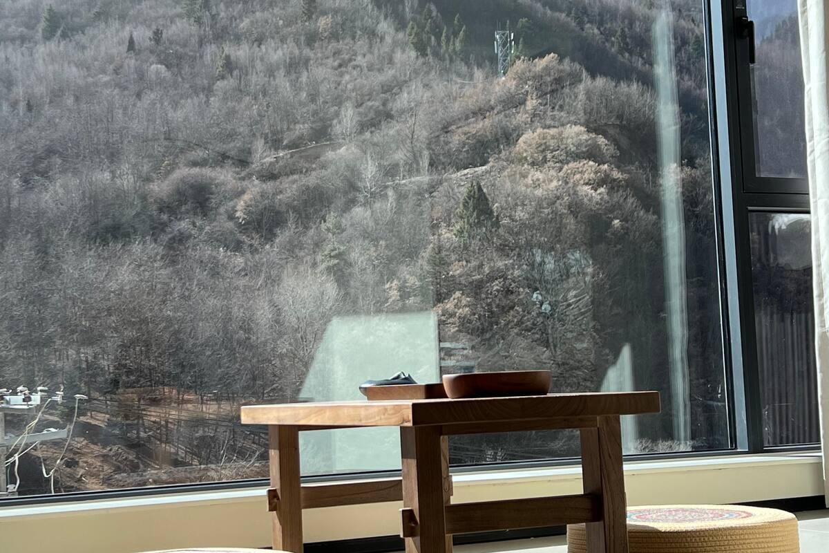 Une table en bois avec un bol et une tasse, une fenêtre donnant sur une colline boisée, et deux petits poufs ronds posés sur le sol.