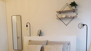 Uma cama bem arrumada com roupa de cama listrada, uma luminária de cabeceira, uma prateleira na parede com uma planta e um espelho grande.