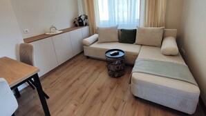 Um espaço de estar compacto com sofá, mesa redonda pequena, uma cadeira e uma janela com cortinas.
