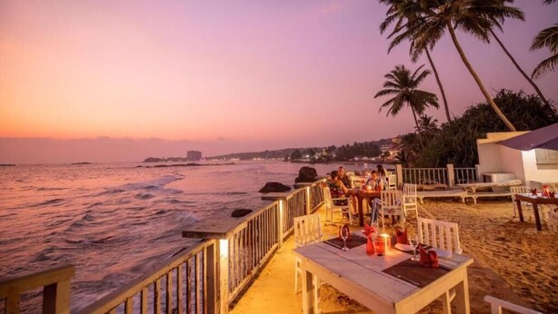 A beachside restaurant with outdoor seating overlooking the ocean, palm trees, and a sunset view. - Rock Fort Beach Resort (Unawatuna)